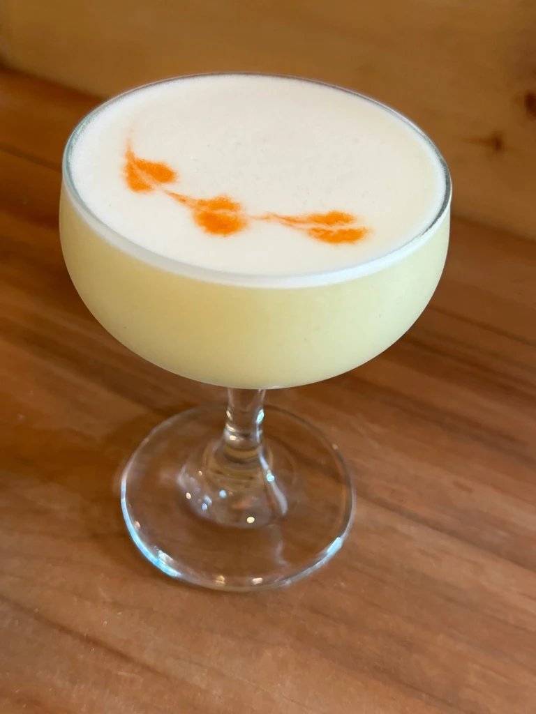 Pale yellow cocktail in coupe glass with orange art on white foam.