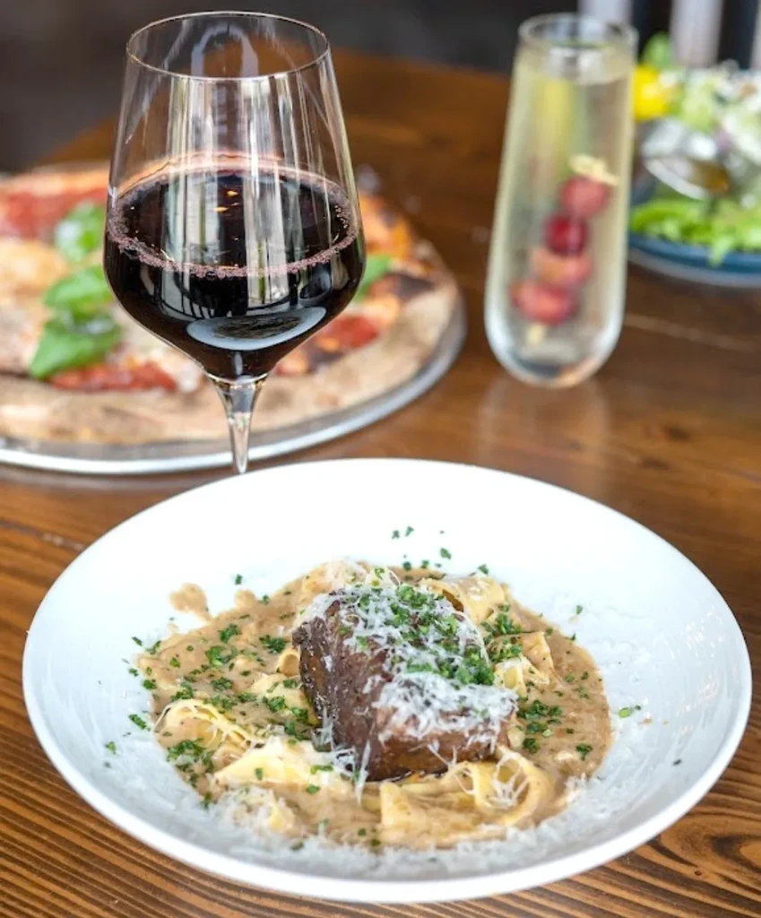 Gourmet pasta with braised meat, red wine, and pizza on a wooden table.