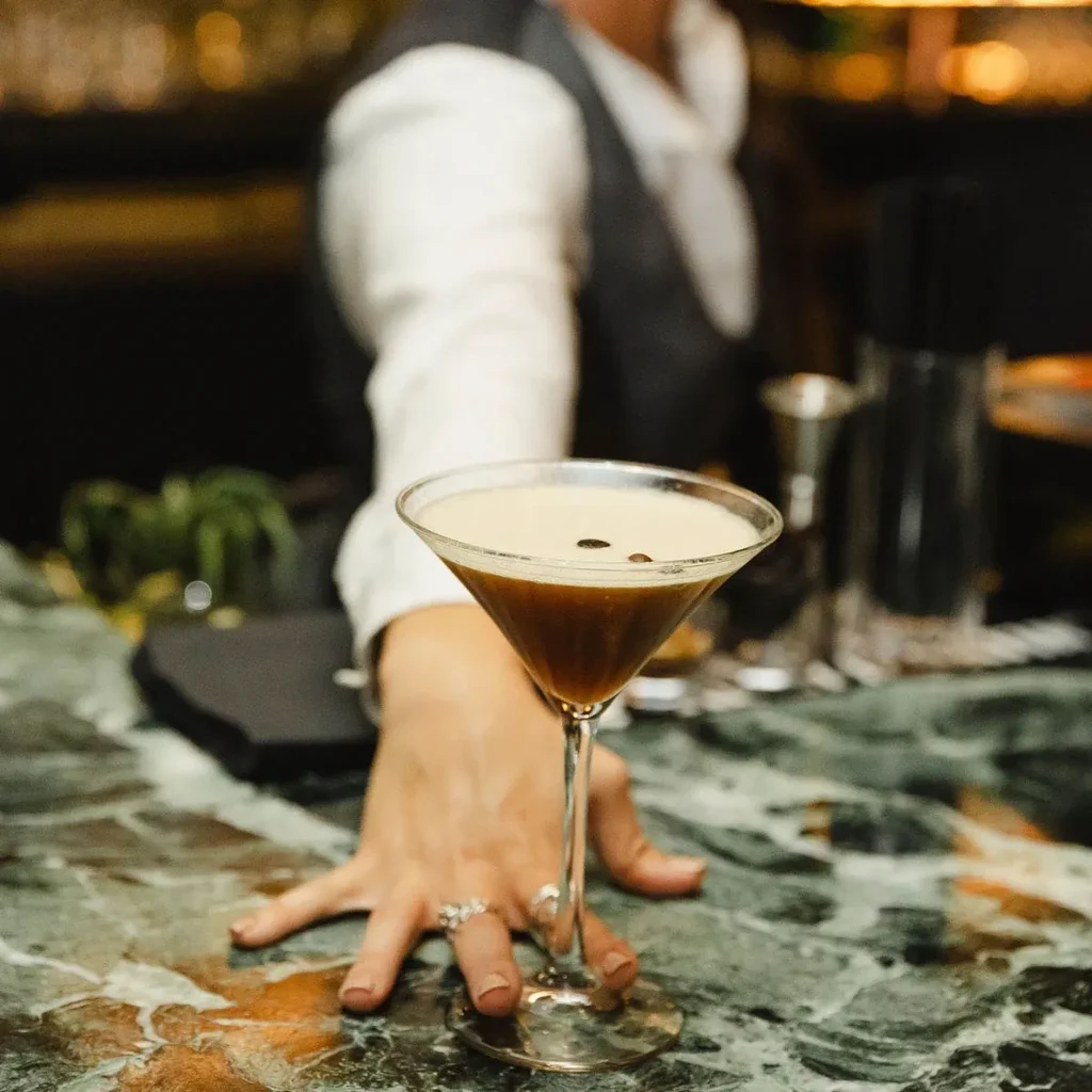 Espresso martini with frothy top, coffee beans, hand on marble bar.