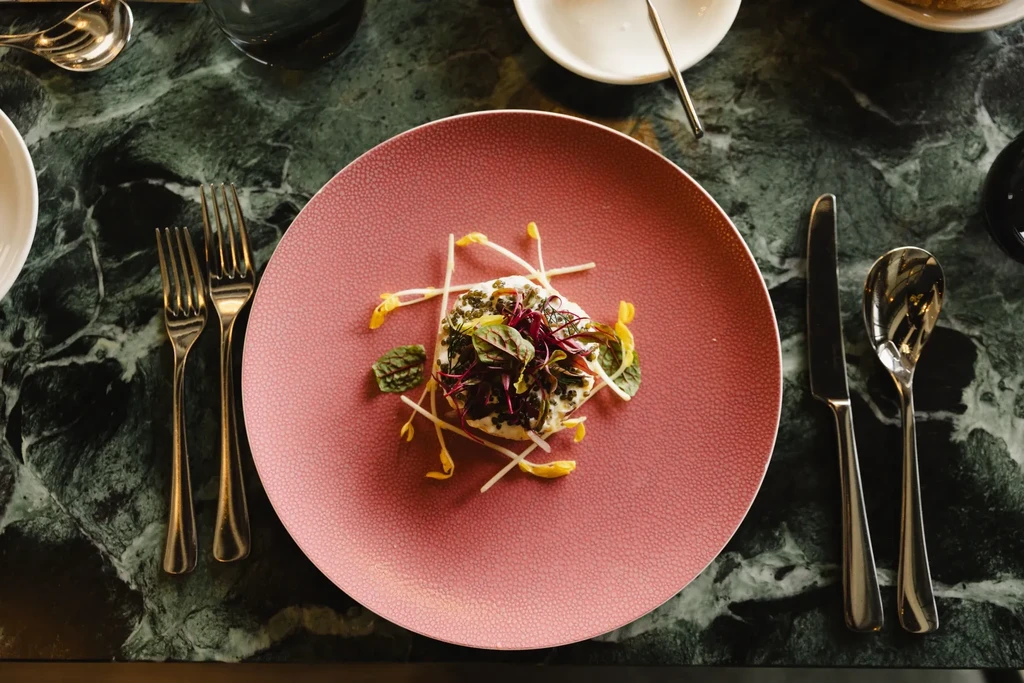 Top-down: Creamy dish with microgreens, pearls, pink plate, marble table.