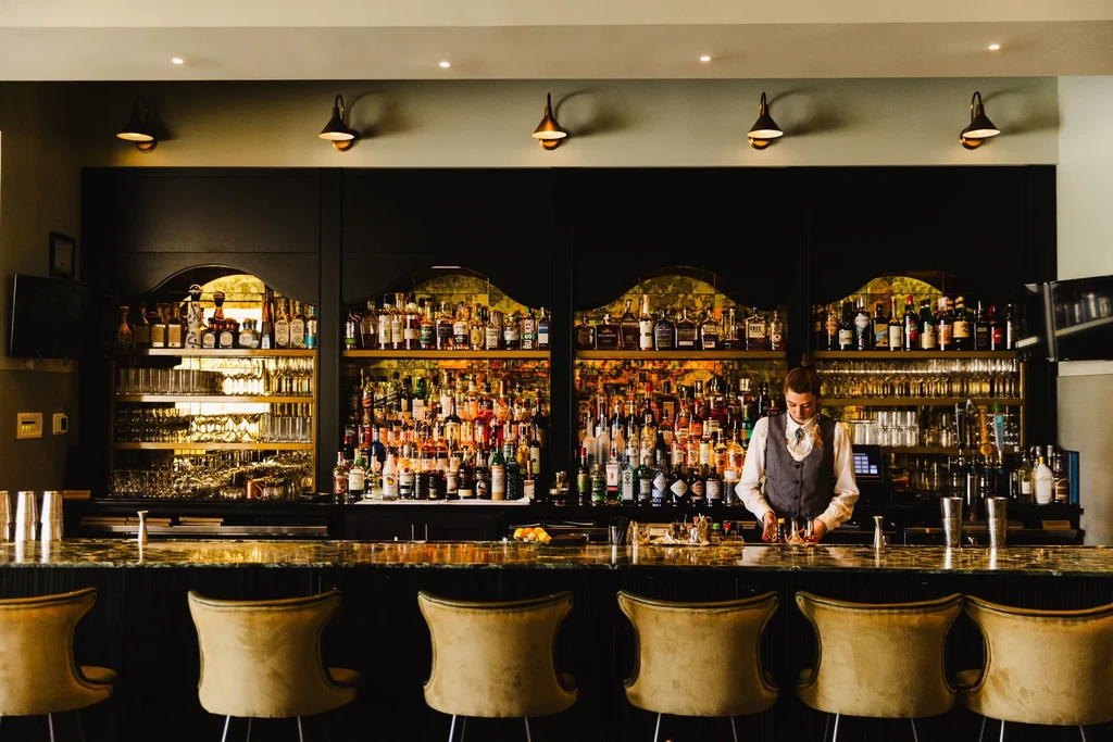 Professional bar interior with bartender, marble counter, liquor bottles, and stools.