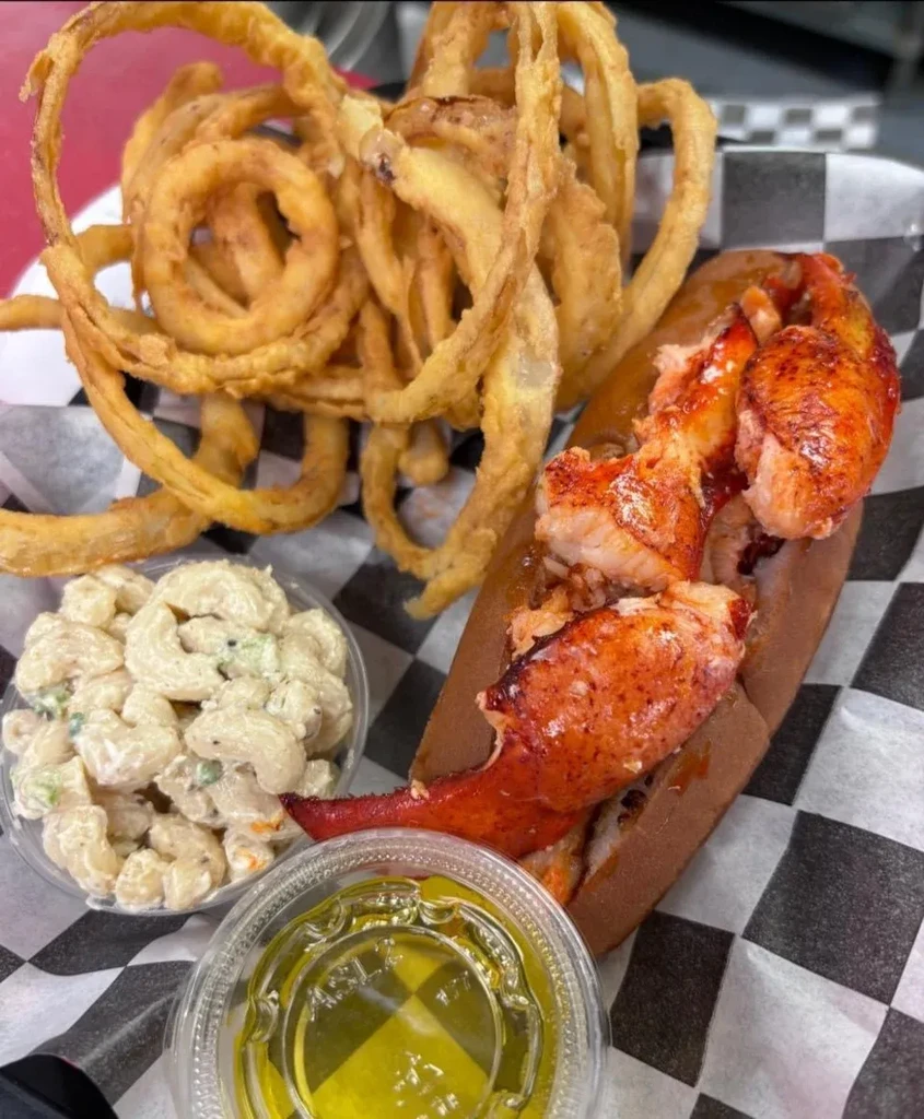 Close-up overhead of a lobster roll, curly fries, macaroni salad, dipping sauce.