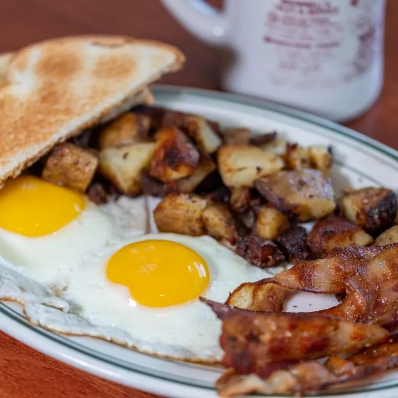 Restaurant plate with sunny-side-up eggs, bacon, roasted potatoes, and toast.