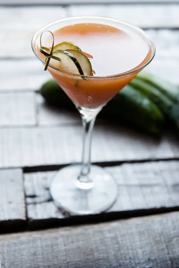 Cold orange cocktail in martini glass, cucumber garnish, rustic table.