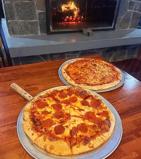 Two freshly baked pizzas, pepperoni and cheese, by a warm fireplace.