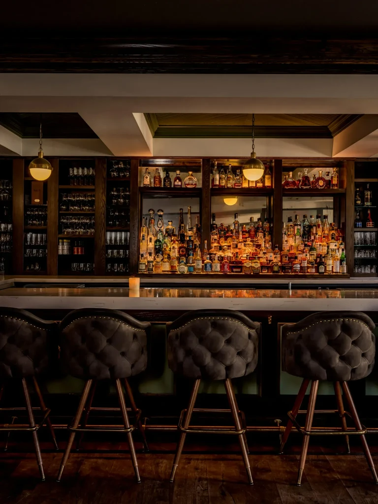 Upscale bar with illuminated liquor bottles and three tufted stools.