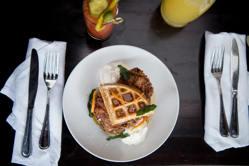 Overhead flat-lay of fried chicken and waffle, garnished, with drinks.