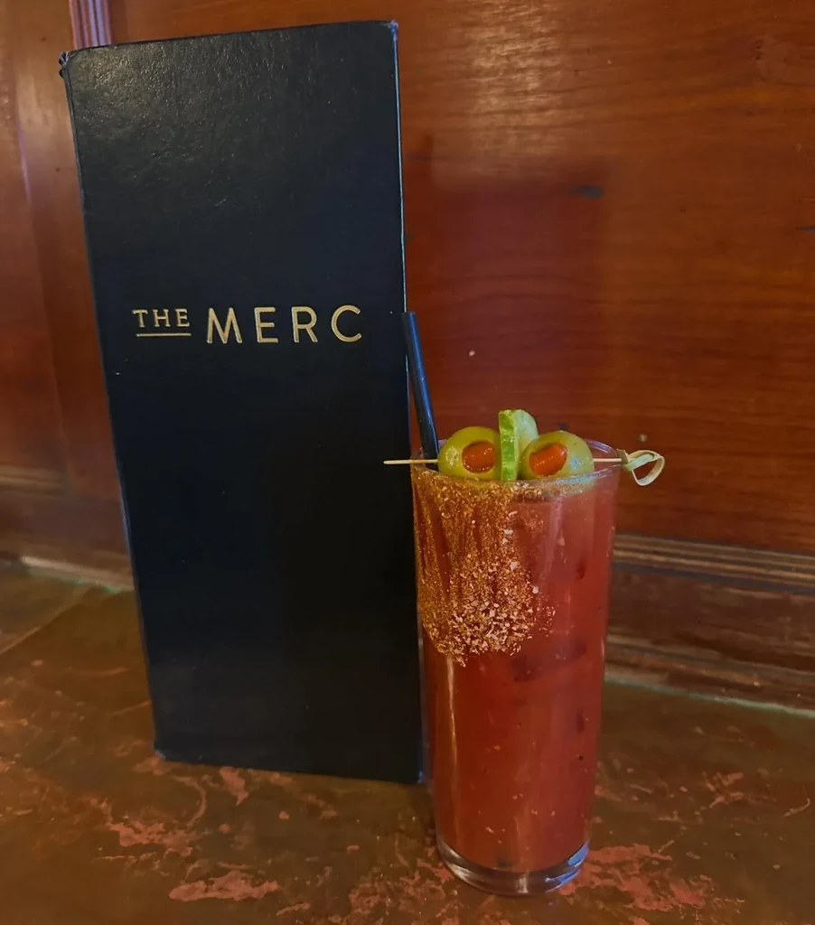 Tall reddish-brown cocktail, olives, salted rim, and 'THE MERC' menu.