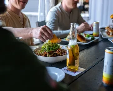 Hand dips chip into nachos; beer bottle and people dining.