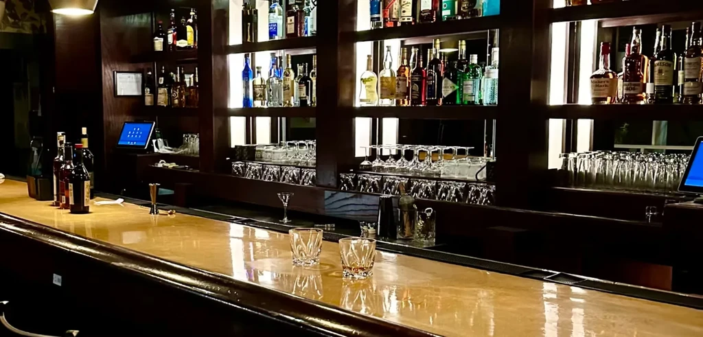 Empty, polished bar counter displaying whiskey glasses and many liquor bottles.