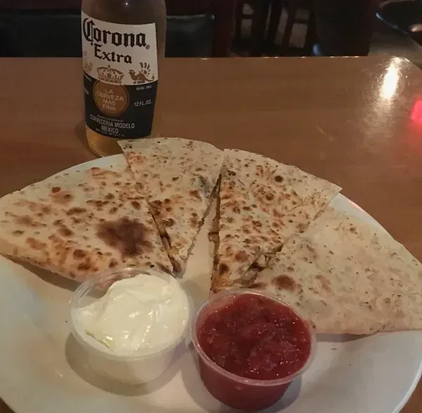 Cut quesadilla, two dips, and Corona beer on a wooden table.