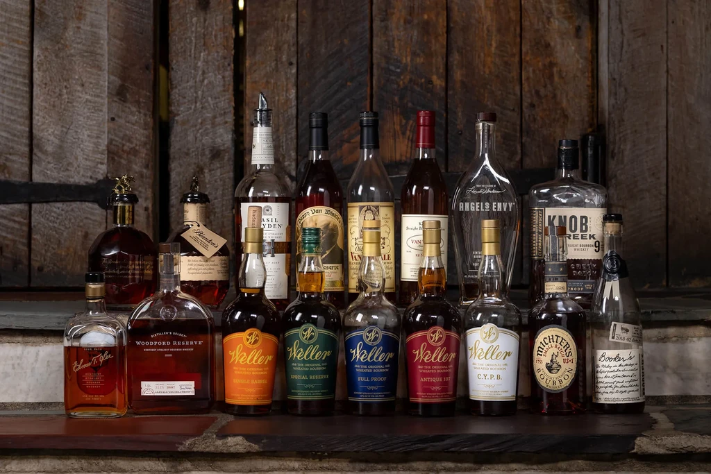 Approximately 20 whiskey and bourbon bottles on a stone and wood surface.