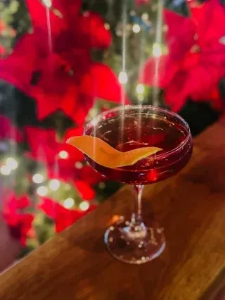 Dark red cocktail with orange peel, blurred poinsettias and fairy lights.