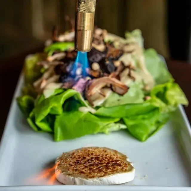 Culinary torch caramelizes cheese slice, flaming a green salad.
