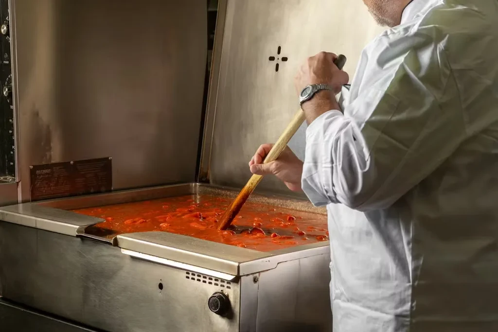 Chef stirs large vat of chunky tomato sauce with wooden spoon.