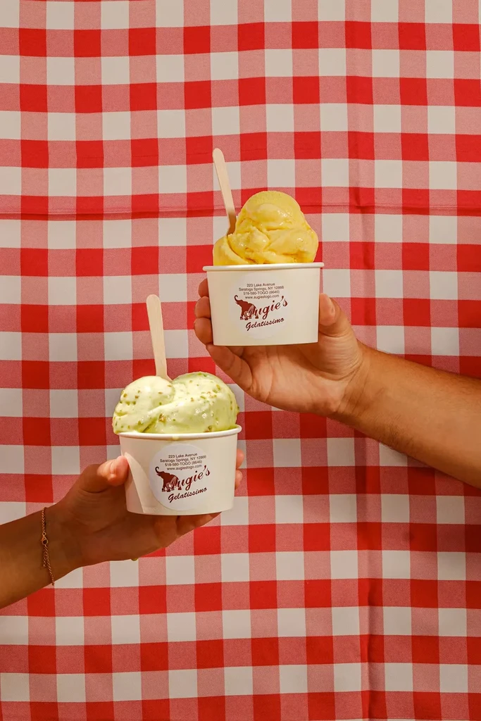Two hands hold Augie's Gelatissimo yellow and green gelato with spoons.
