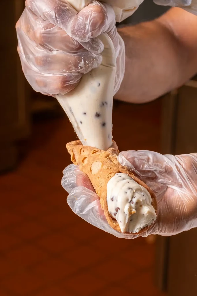 Gloved hands pipe chocolate chip cream into a crispy cannoli shell.