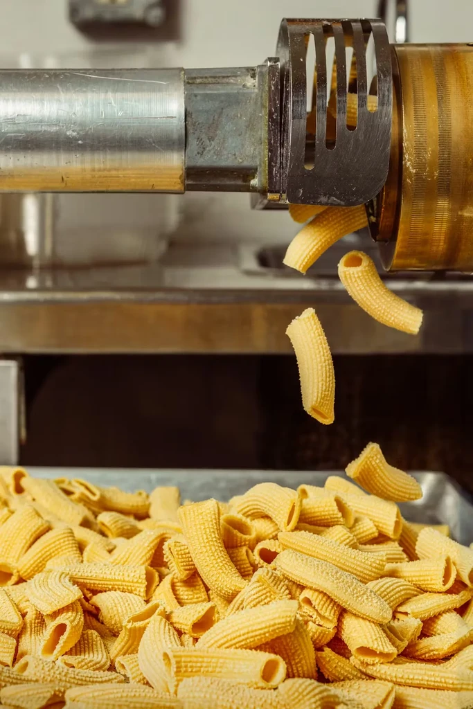 Golden-yellow rigatoni pasta extruding from a machine into a tray.
