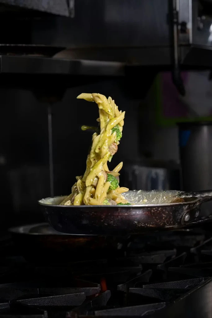 Green penne pasta with broccoli tossed mid-air from a dark pan.