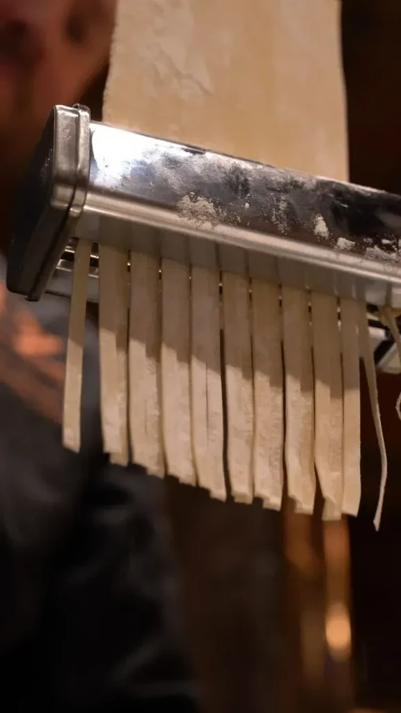 Stainless steel pasta maker cutting fresh tagliatelle from light-colored dough.