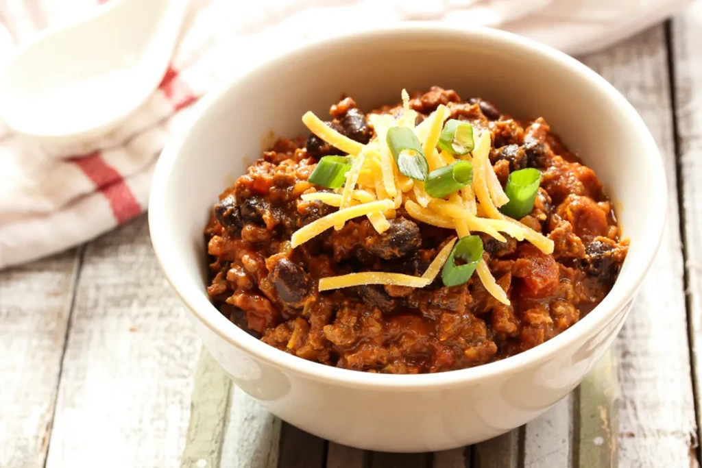 Hearty chili in a white bowl, topped with cheese and green onions.