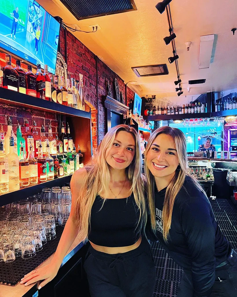 Two young smiling women in a bar with bottles, glasses, and TVs.