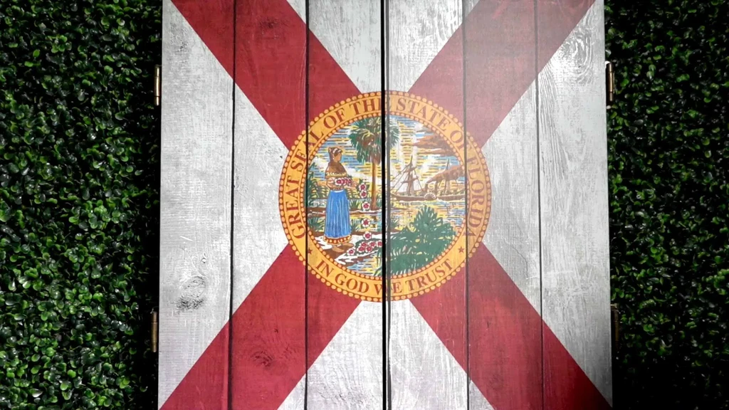 Wooden bi-fold panel painted with Florida flag, surrounded by green hedge.