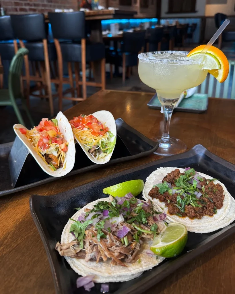 Close-up of four tacos and a margarita in a modern restaurant setting.