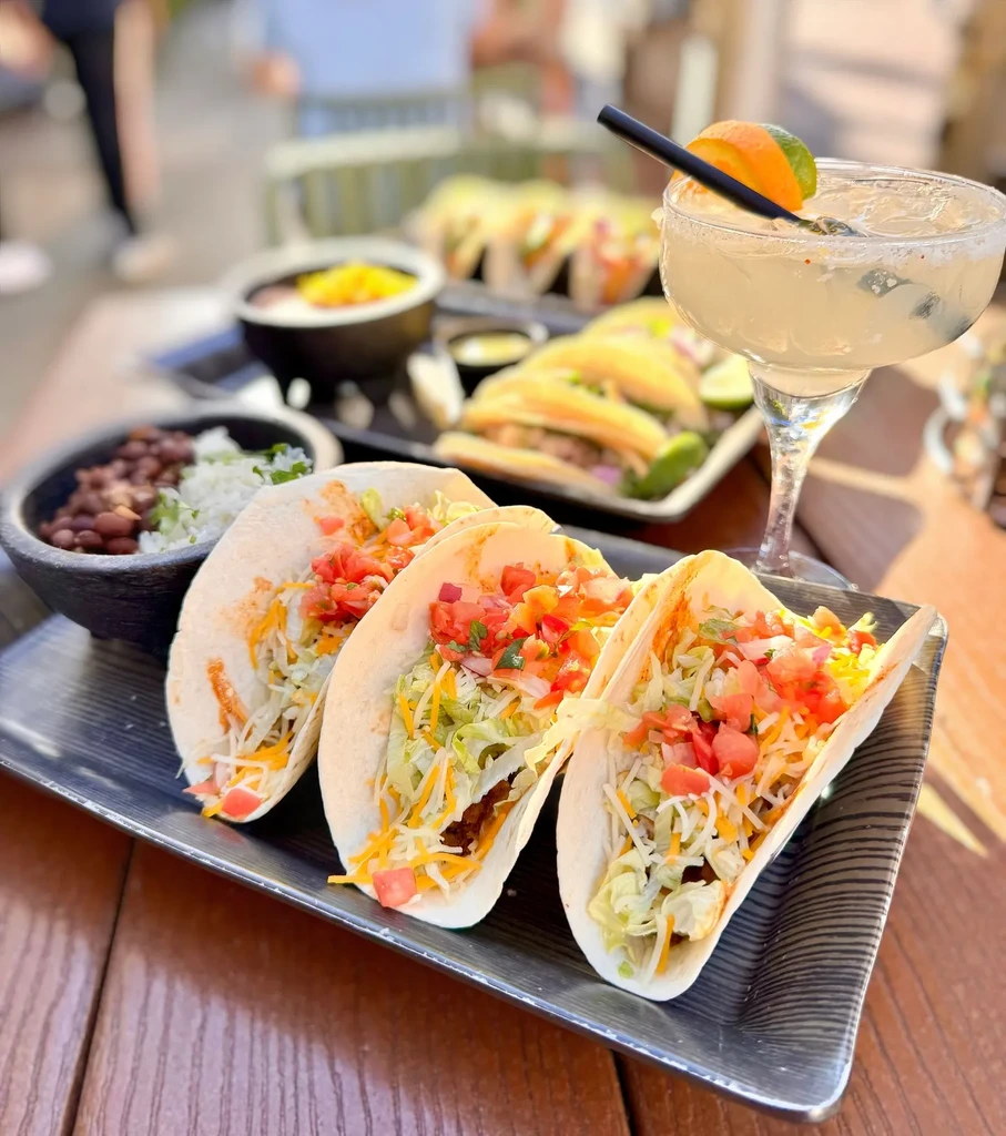 Three soft-shell tacos, margarita, rice, and beans on a wooden table.