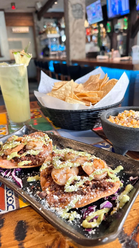 Two shrimp tostadas, Esquites, a drink, and chips in a bar.
