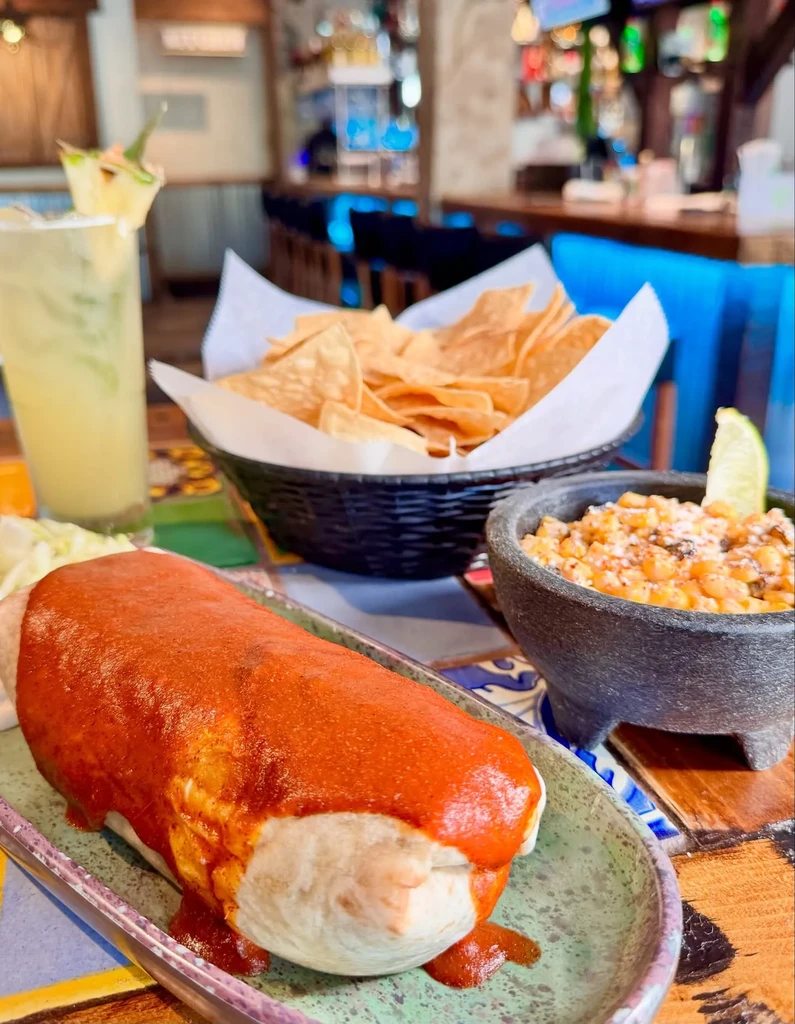 Generously sauced burrito, corn salad, and drink on restaurant table.