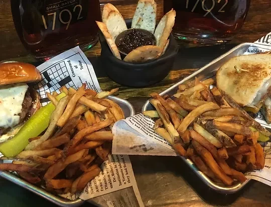 Overhead: Burger, grilled sandwich, fries, and bourbon on a rustic table.