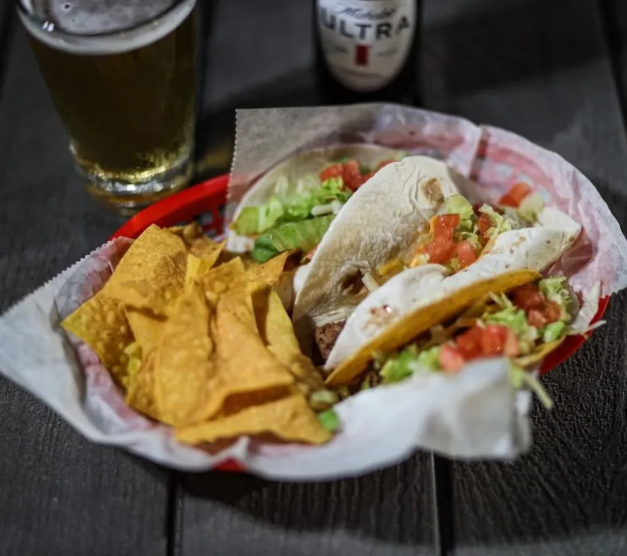 Three tacos, chips, guacamole in a basket with beer on a wooden table.