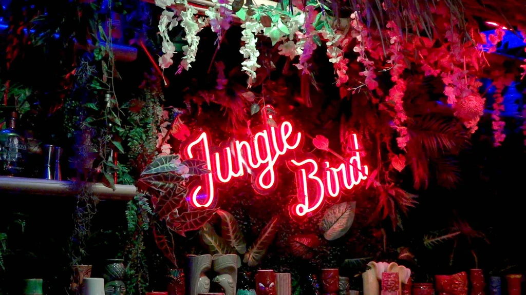 Red neon "Jungle Bird" sign in a dark, foliage-filled tiki bar.