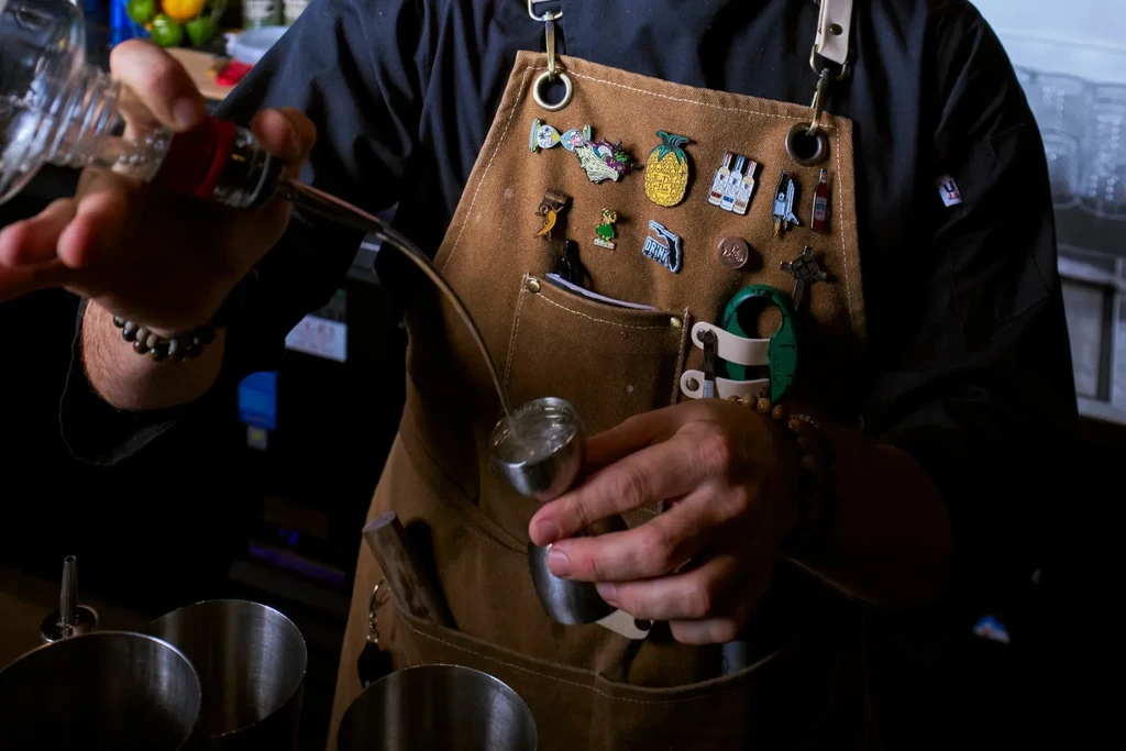 Person's hands pouring liquid into a jigger, wearing a pin-covered apron.
