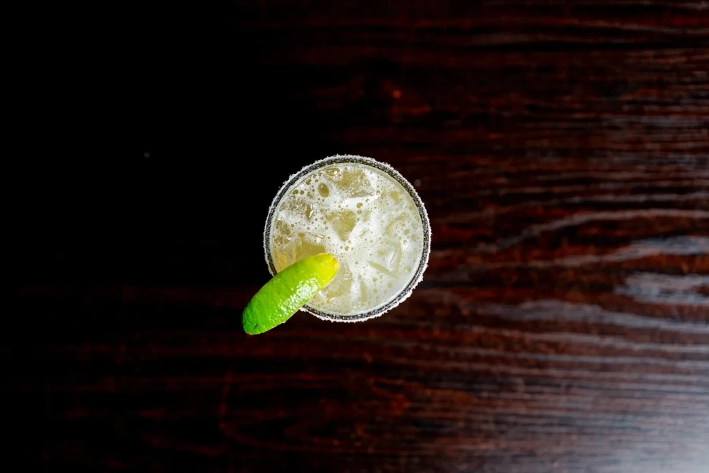 Overhead margarita with lime, salted rim, and ice on dark wood.