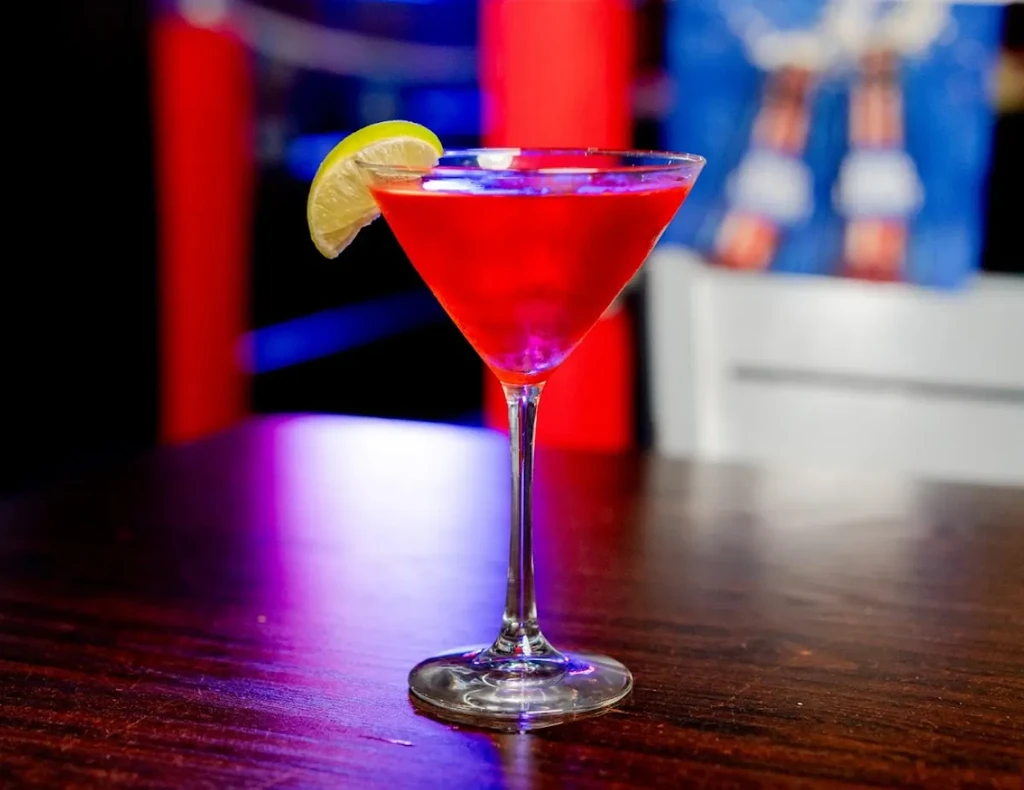 Neon red cocktail in a martini glass with lime on dark wood.