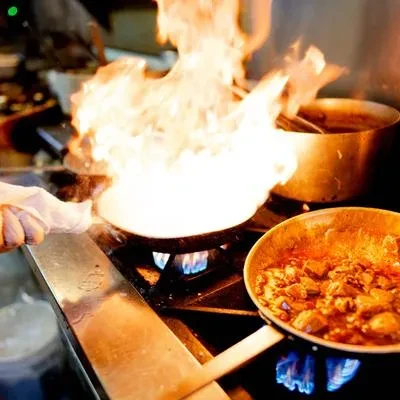 Professional kitchen stovetop with one pan flambéing, another cooking stew.