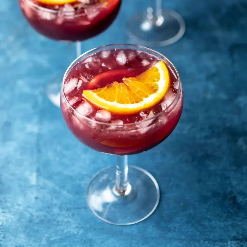Vibrant red-purple drink with ice, orange slice, and submerged fruit.