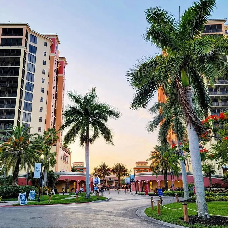 Warmly lit tropical street with tall buildings, palms, and strolling people.