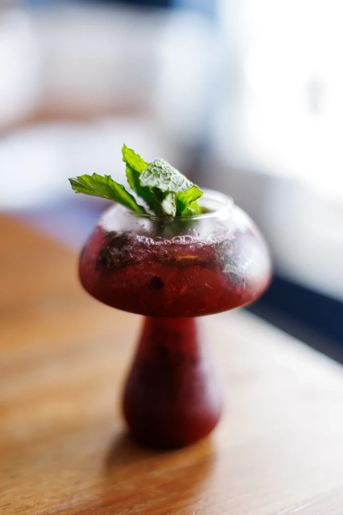 Red drink with mint in a mushroom-shaped glass on a wooden table.