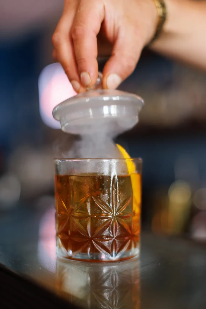 Hand lifts lid from smoked cocktail, white smoke rising.