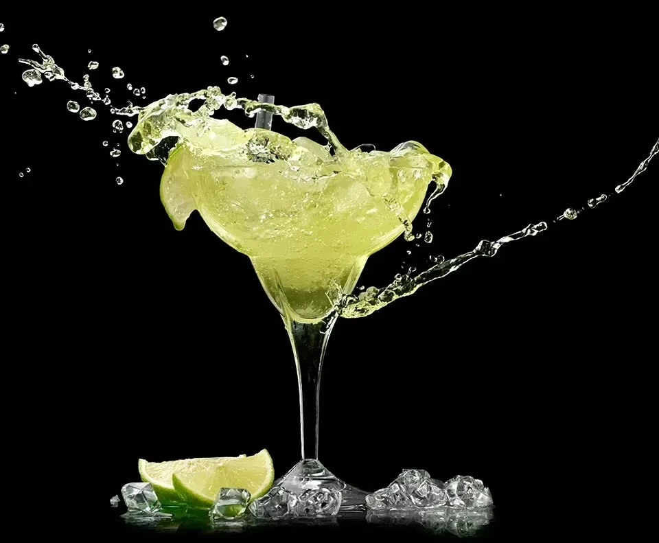 Overflowing green-yellow margarita splashing with ice and lime wedges.