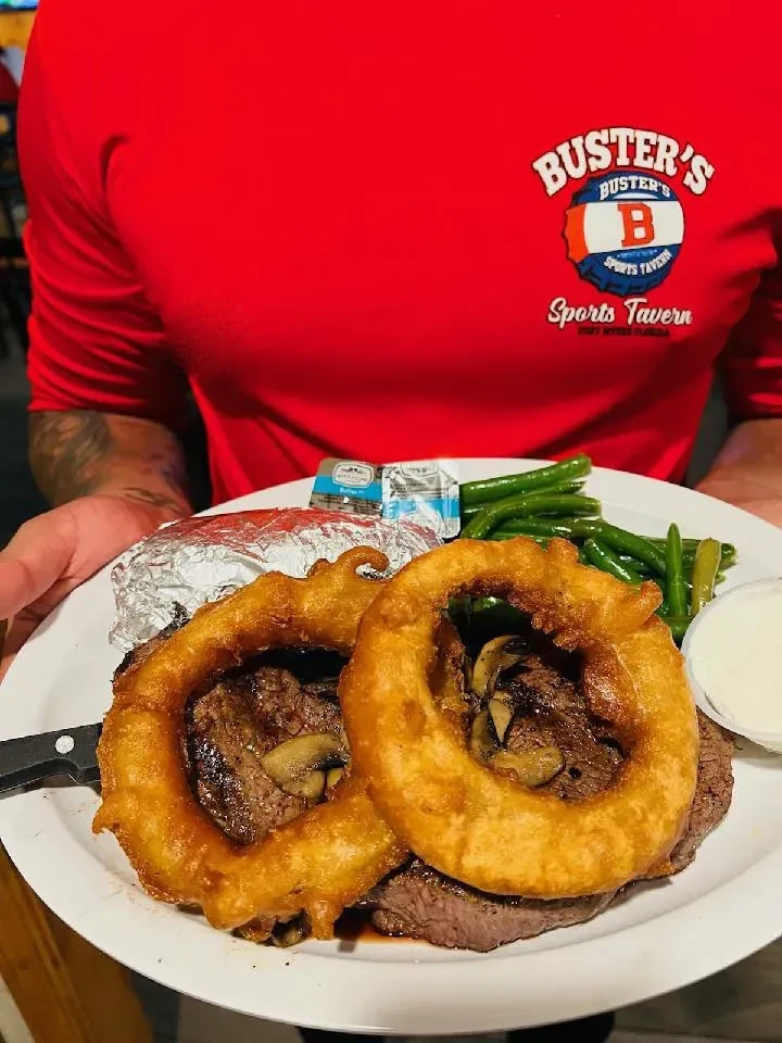 Person holds plate with steak, mushrooms, onion rings, beans, and potato.