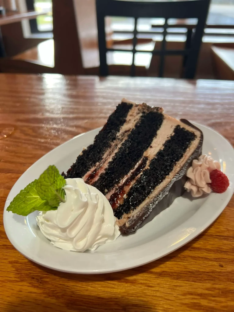 Multilayered chocolate cake slice with cream, raspberry, mint, on table.