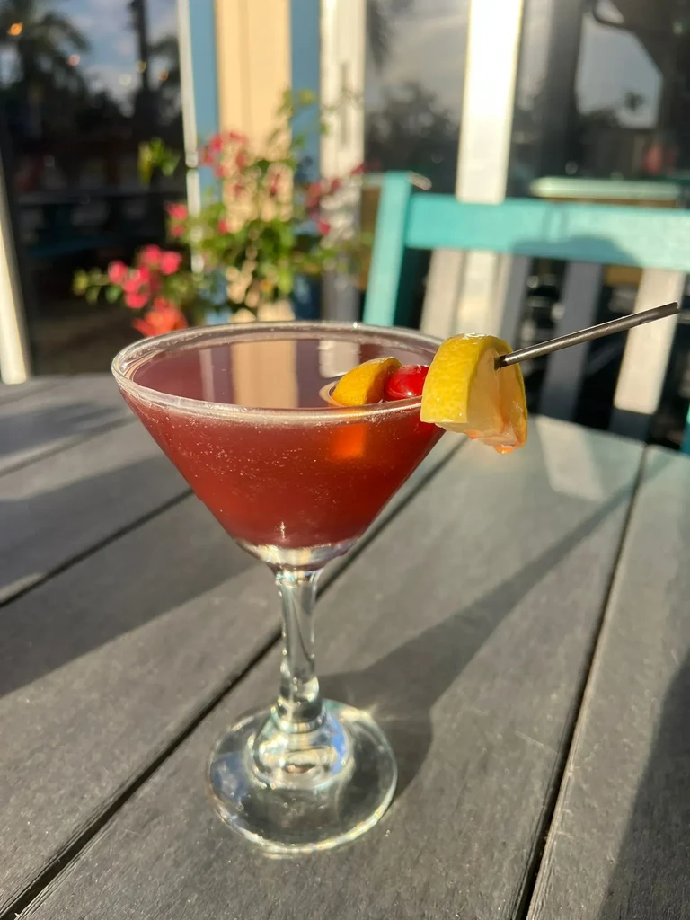Deep red cocktail in martini glass, with skewered lemon twist and cherry.