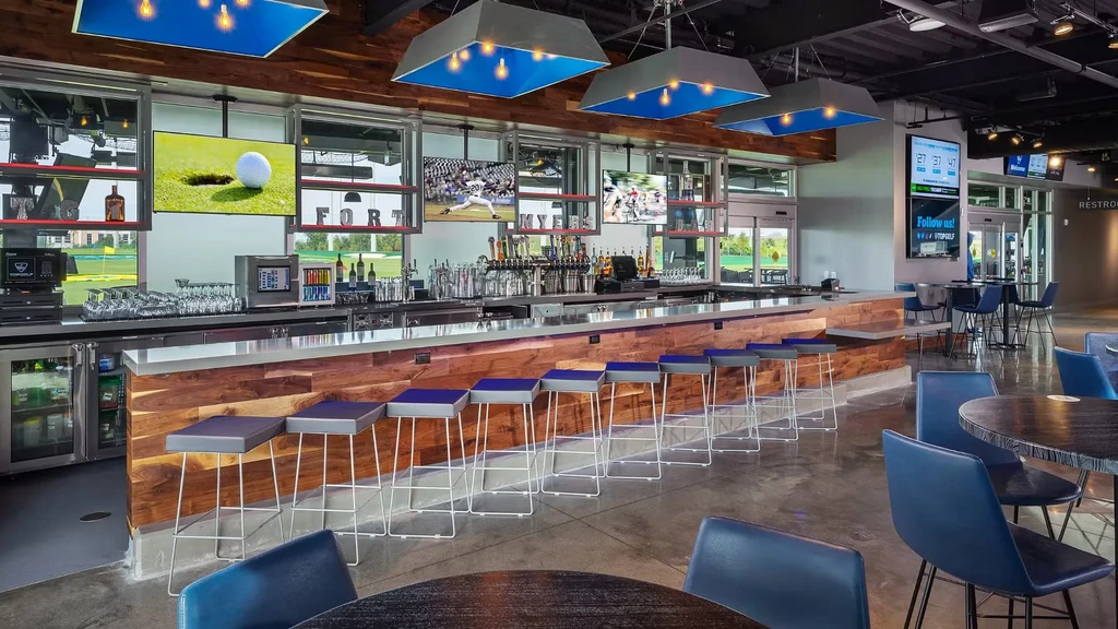 Modern Topgolf bar with sports TVs, blue stools, and outdoor golf range.