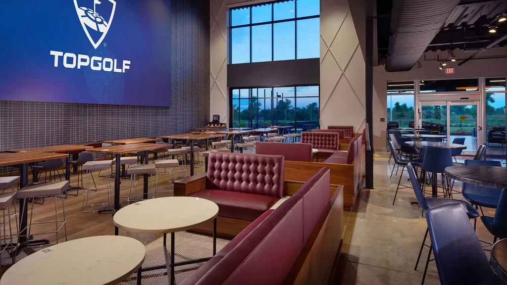 Contemporary Topgolf dining lounge interior with logo screen, varied seating, windows.