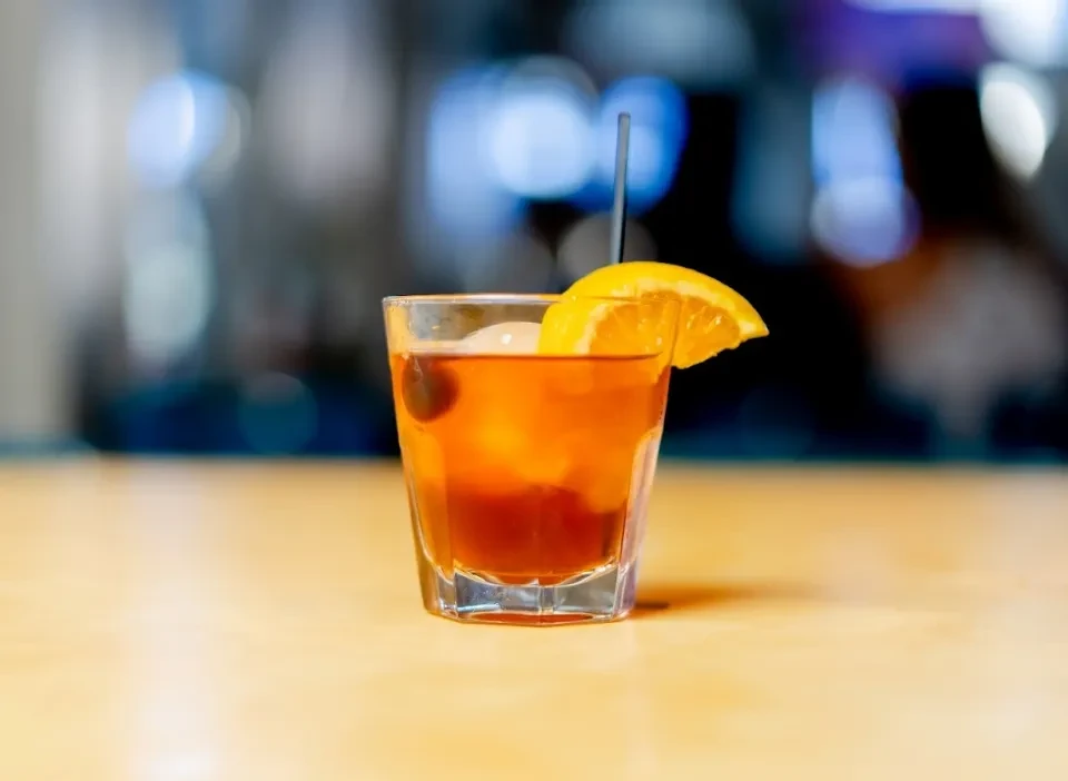 Amber cocktail in faceted glass with ice, orange slice, and dark garnishes.