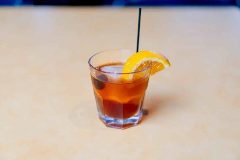 Amber cocktail with ice, cherries, orange slice, and straw in faceted glass.